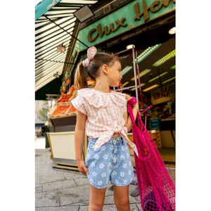 A young girl wearing a striped top and the Louis Louise Girls’ Andrea Denim Floral Short stands outside a store, holding a pink net bag and looking to the side.