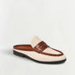 The Dean Canvas Como Loafer Mule from Simkhai features a cream-colored canvas upper with brown leather strap and trim, a black sole, and stitched detailing. This stylish backless loafer is displayed on a plain white background.