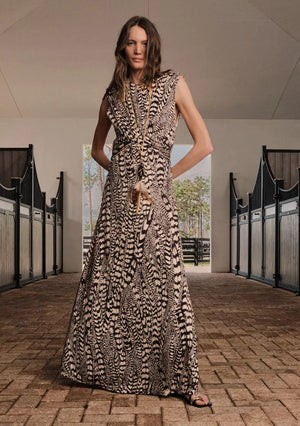 A woman stands in a stable hallway wearing the ankle-length Silent Rising Dress by Johanna Ortiz, crafted from double-sided jersey and adorned with an elegant black and white External Feathers motif, complemented by a long necklace.