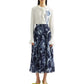 A woman wears Erdem's Botanical Engraving Silk Pleated Midi Skirt, a navy piece adorned with a delicate white floral print, paired with a white blouse featuring ruffled collar and cuffs.
