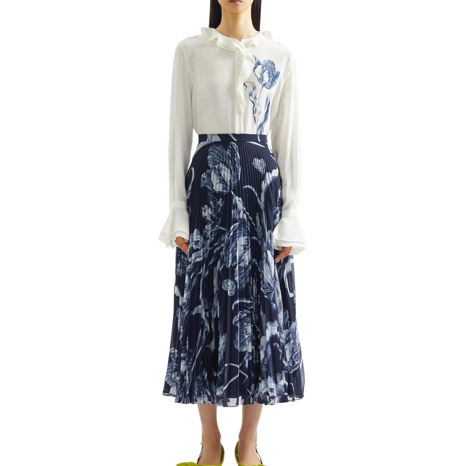 A woman wears Erdem's Botanical Engraving Silk Pleated Midi Skirt, a navy piece adorned with a delicate white floral print, paired with a white blouse featuring ruffled collar and cuffs.