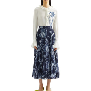 A woman wears Erdem's Botanical Engraving Silk Pleated Midi Skirt, a navy piece adorned with a delicate white floral print, paired with a white blouse featuring ruffled collar and cuffs.