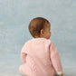 A baby wearing the Louise Misha Baby Girls’ Drilia Overalls in pink, featuring scalloped finishes, sits on the floor facing away from the camera against a light blue background.
