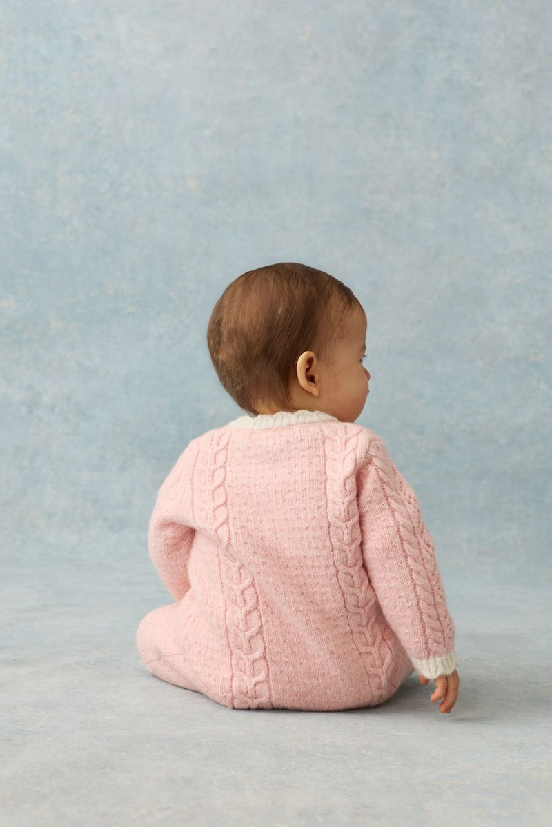 A baby wearing the Louise Misha Baby Girls’ Drilia Overalls in pink, featuring scalloped finishes, sits on the floor facing away from the camera against a light blue background.