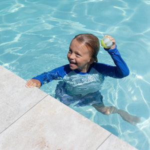 A child in a blue swimsuit stands by the pool’s edge, smiling and ready for water play, while holding a Sunnylife Tides Out Citrus Skim Ball from the set.
