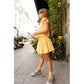A young girl wearing the Louis Louise Girls’ Opera Skirt and sunglasses stands on a city sidewalk holding a stick, with plants and buildings in the background.