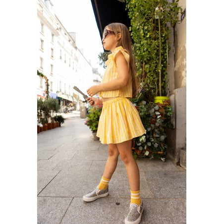 A young girl wearing the Louis Louise Girls’ Opera Skirt and sunglasses stands on a city sidewalk holding a stick, with plants and buildings in the background.