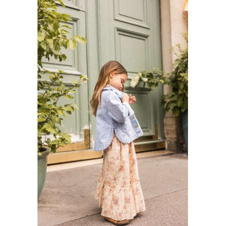 A young girl wearing the Louis Louise Girls’ Rachel Skirt and a denim jacket stands on a sidewalk in front of a large green door, looking down at something in her hands.