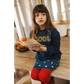 A young girl with long brown hair sits on a wooden table, holding an open book. She wears a “COOL” sweater and the Louis Louise Girls’ Teacher Skirt, an A-line denim skirt for girls, paired with vibrant red tights. A bowl of fruit is in the background.