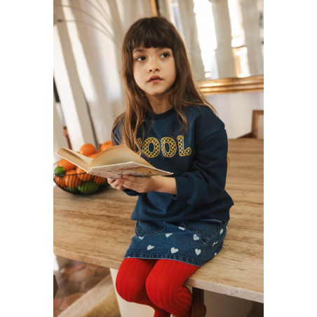 A young girl with long brown hair sits on a wooden table, holding an open book. She wears a “COOL” sweater and the Louis Louise Girls’ Teacher Skirt, an A-line denim skirt for girls, paired with vibrant red tights. A bowl of fruit is in the background.