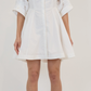 A woman stands against a plain background wearing the Simkhai Jazzie Mini Dress, a white poplin button-down style with rolled-up sleeves, paired with white high-heeled sandals.