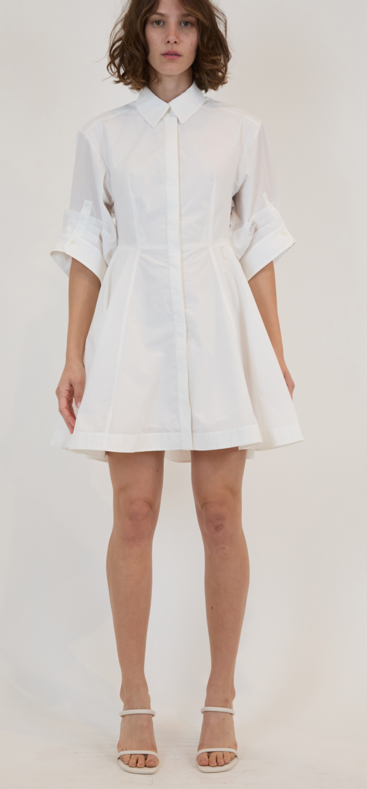 A woman stands against a plain background wearing the Simkhai Jazzie Mini Dress, a white poplin button-down style with rolled-up sleeves, paired with white high-heeled sandals.