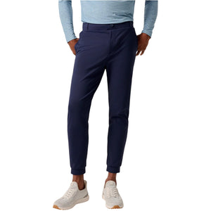 A person wearing a light blue long-sleeve shirt, Johnnie-O Solom Pant men's joggers in dark blue stretch fabric, and white sneakers stands with hands in pockets against a white background.