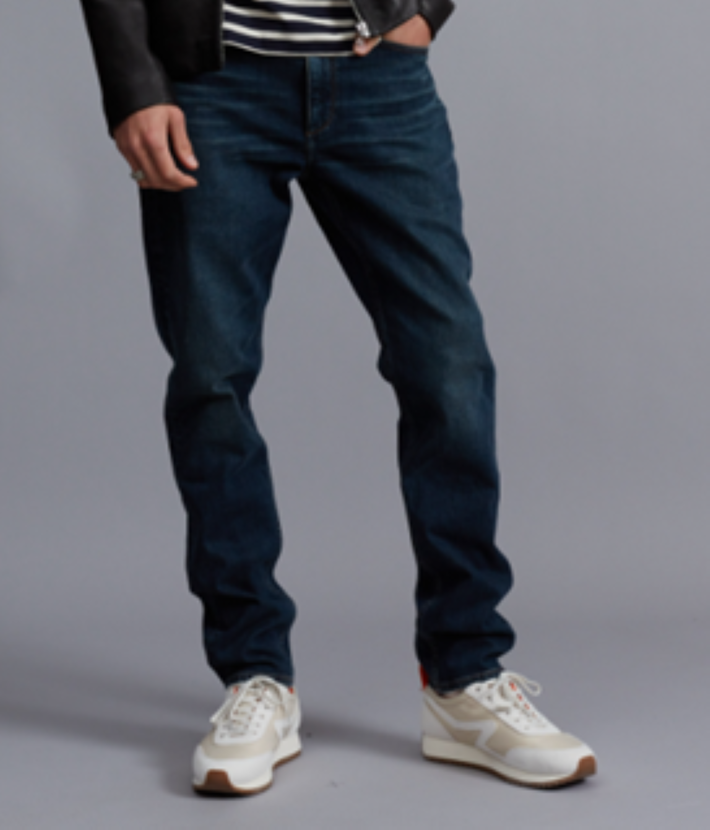 A person wearing Rag & Bone Fit 3 Authentic Stretch dark blue athletic fit jeans, a black jacket, and white sneakers stands against a plain gray background.