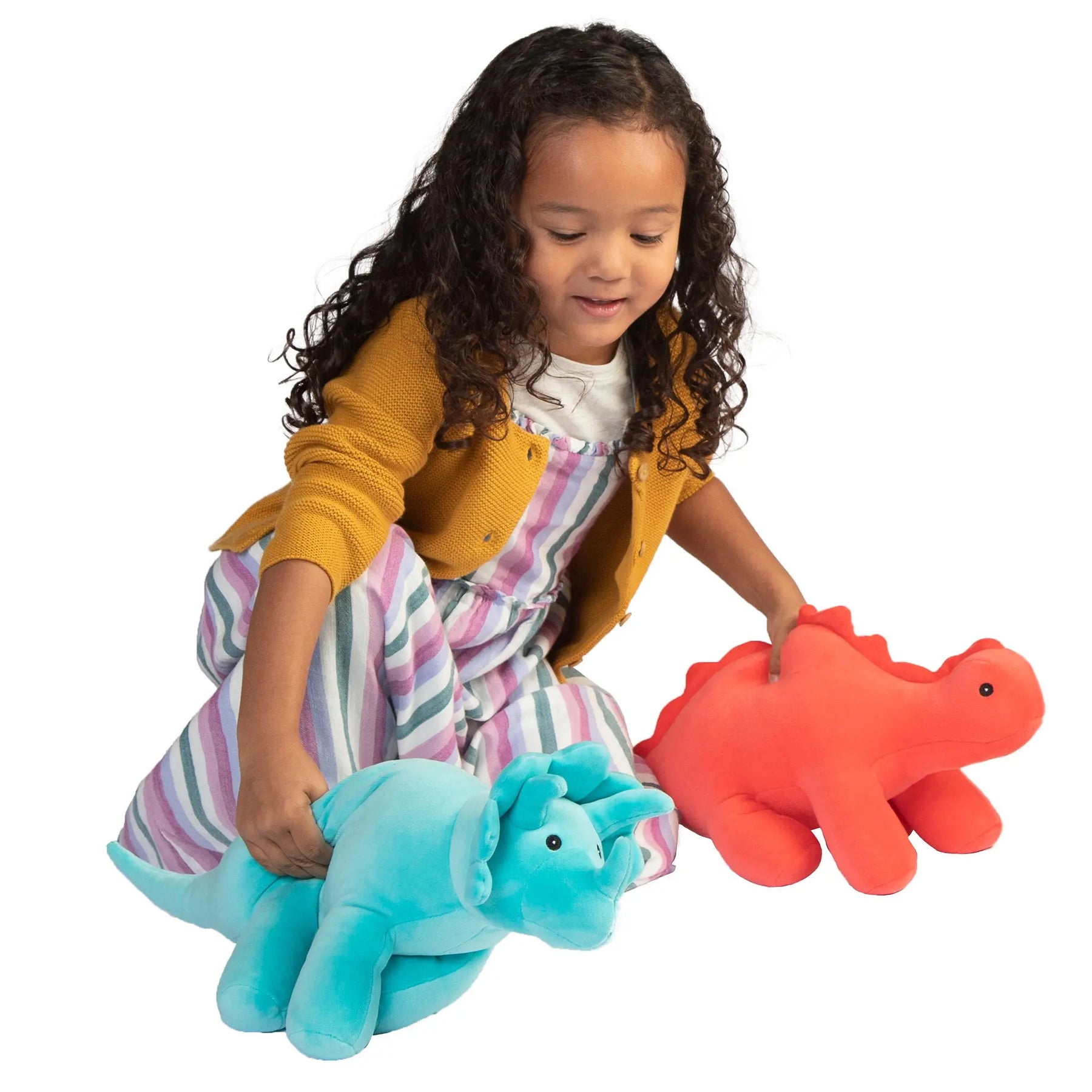 A young girl with curly hair is playing with a blue Manhattan Toy Velveteen Dino plush toy and an orange Manhattan Toy Triceratops plush toy, while wearing a striped dress and a yellow cardigan. These are some of the Best Toys from Toy Fair 2020.