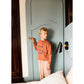 A young boy wearing an orange sweatshirt and Arsene & Les Pipelettes Boys' Striped Sun Lounger Pants with an elasticated waist stands by a closed blue door, holding the handle, with a black wall lamp and a bed in the foreground.