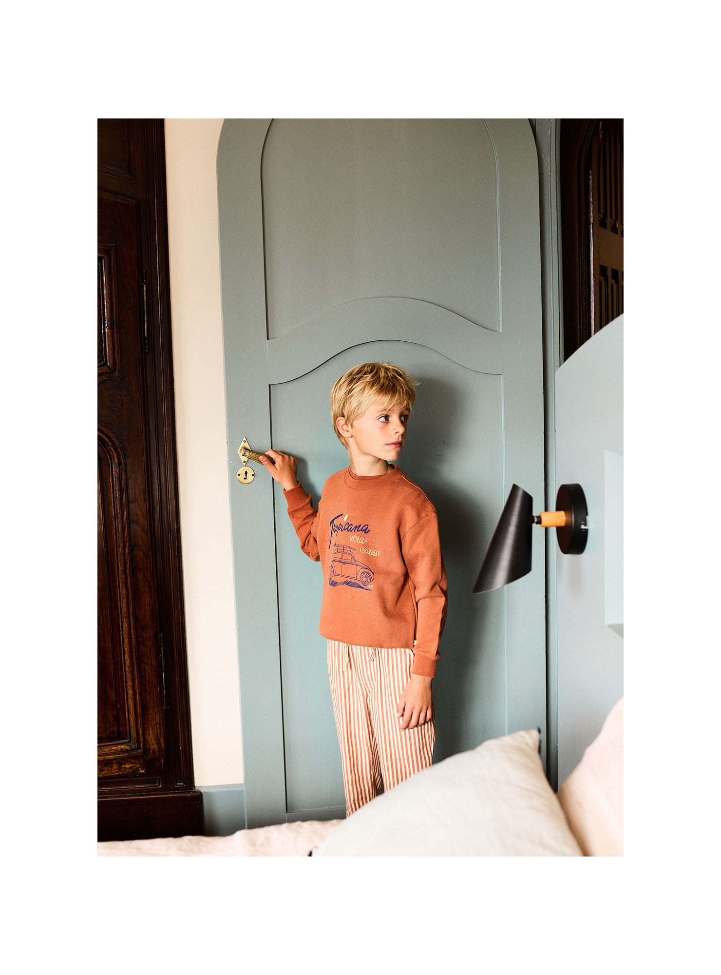 A young boy wearing an orange sweatshirt and Arsene & Les Pipelettes Boys' Striped Sun Lounger Pants with an elasticated waist stands by a closed blue door, holding the handle, with a black wall lamp and a bed in the foreground.