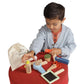 A young boy delights in imaginative role-play using the Manhattan Toy Style & Groom set, which includes wooden grooming toys such as a comb, scissors, mirror, and spray bottle. He sits on a red stool with the "Style & Groom" canvas bag from Manhattan Toy by his side, filled with his favorite grooming playset for kids.