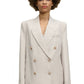 A person with an afro hairstyle is wearing the Dorothee Schumacher Summer Cruise Jacket, a light beige linen blend double-breasted blazer, along with matching trousers, standing against a plain white background. Perfect for summer suiting, this lightweight ensemble from Dorothee Schumacher exudes both style and comfort.