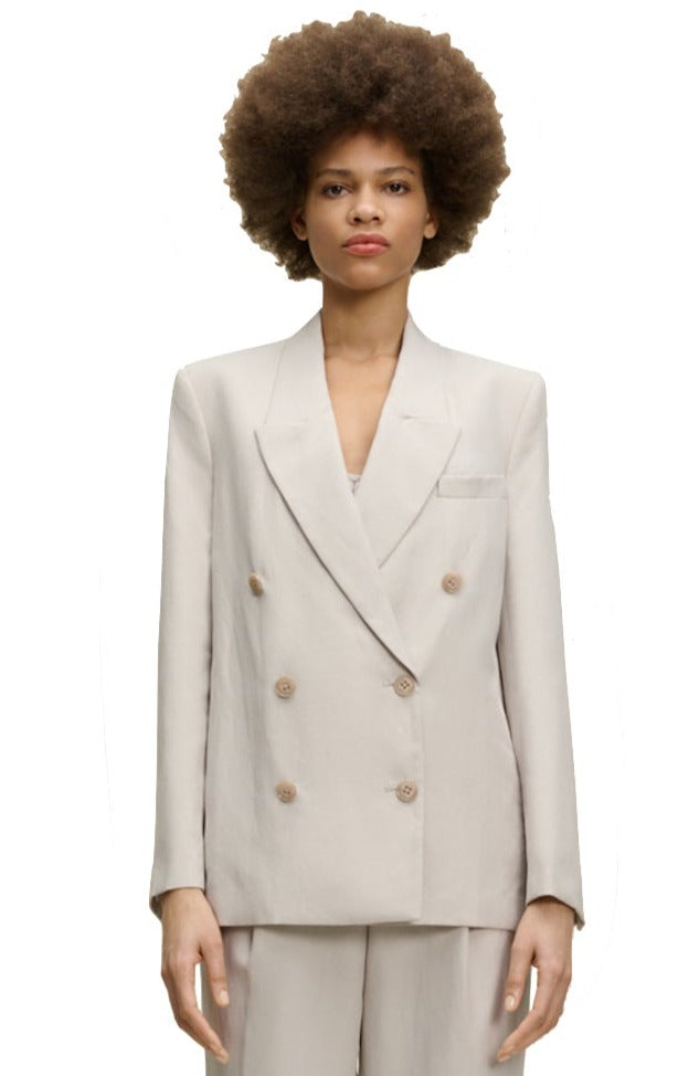 A person with an afro hairstyle is wearing the Dorothee Schumacher Summer Cruise Jacket, a light beige linen blend double-breasted blazer, along with matching trousers, standing against a plain white background. Perfect for summer suiting, this lightweight ensemble from Dorothee Schumacher exudes both style and comfort.