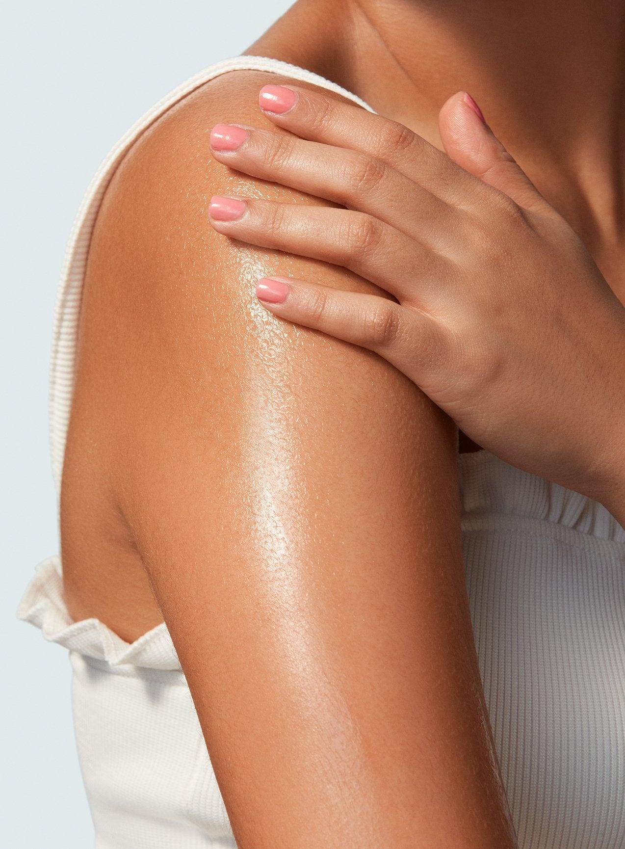 Close-up of a person applying Supergoop! Glow Oil SPF 50 to their shoulder, highlighting moisturized skin and manicured nails.