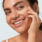 A woman with a radiant smile applying Supergoop! Glowscreen SPF 40, Sunrise liquid foundation with sunscreen on her cheek with a sponge, against a light background.