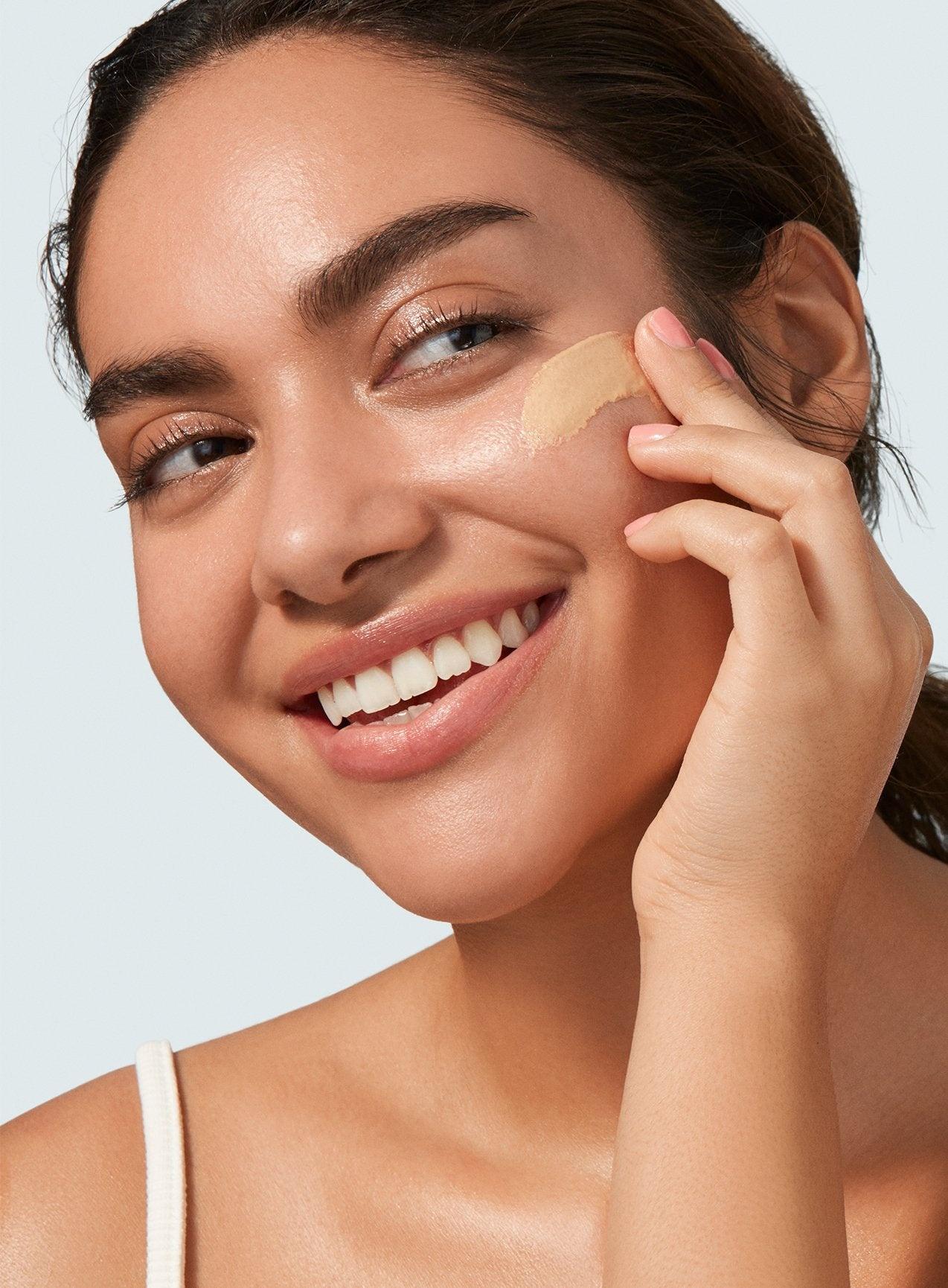 A woman with a radiant smile applying Supergoop! Glowscreen SPF 40, Sunrise liquid foundation with sunscreen on her cheek with a sponge, against a light background.