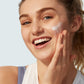 A woman smiles as she applies Supergoop Play Everyday Lotion with Sunflower Extract SPF 50 (2.4 oz) to her cheek, enjoying its gentle, hydrating touch.