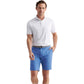 A man wearing a white polo, Peter Millar Surge Performance Short in blue, and a gray belt stands with one hand in his pocket against a white background.