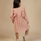 A child with long dark hair, wearing a peach-colored Early Sunday Suzanne Dress adorned with delicate floral embroidery and silver shoes, stands facing away on a beige background.