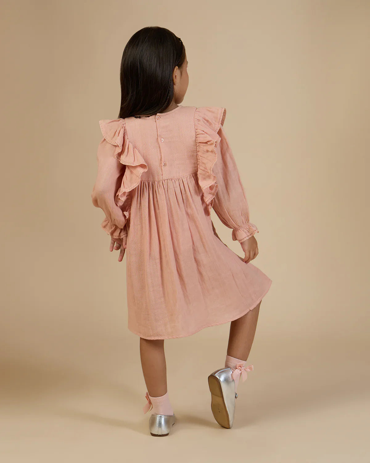 A child with long dark hair, wearing a peach-colored Early Sunday Suzanne Dress adorned with delicate floral embroidery and silver shoes, stands facing away on a beige background.