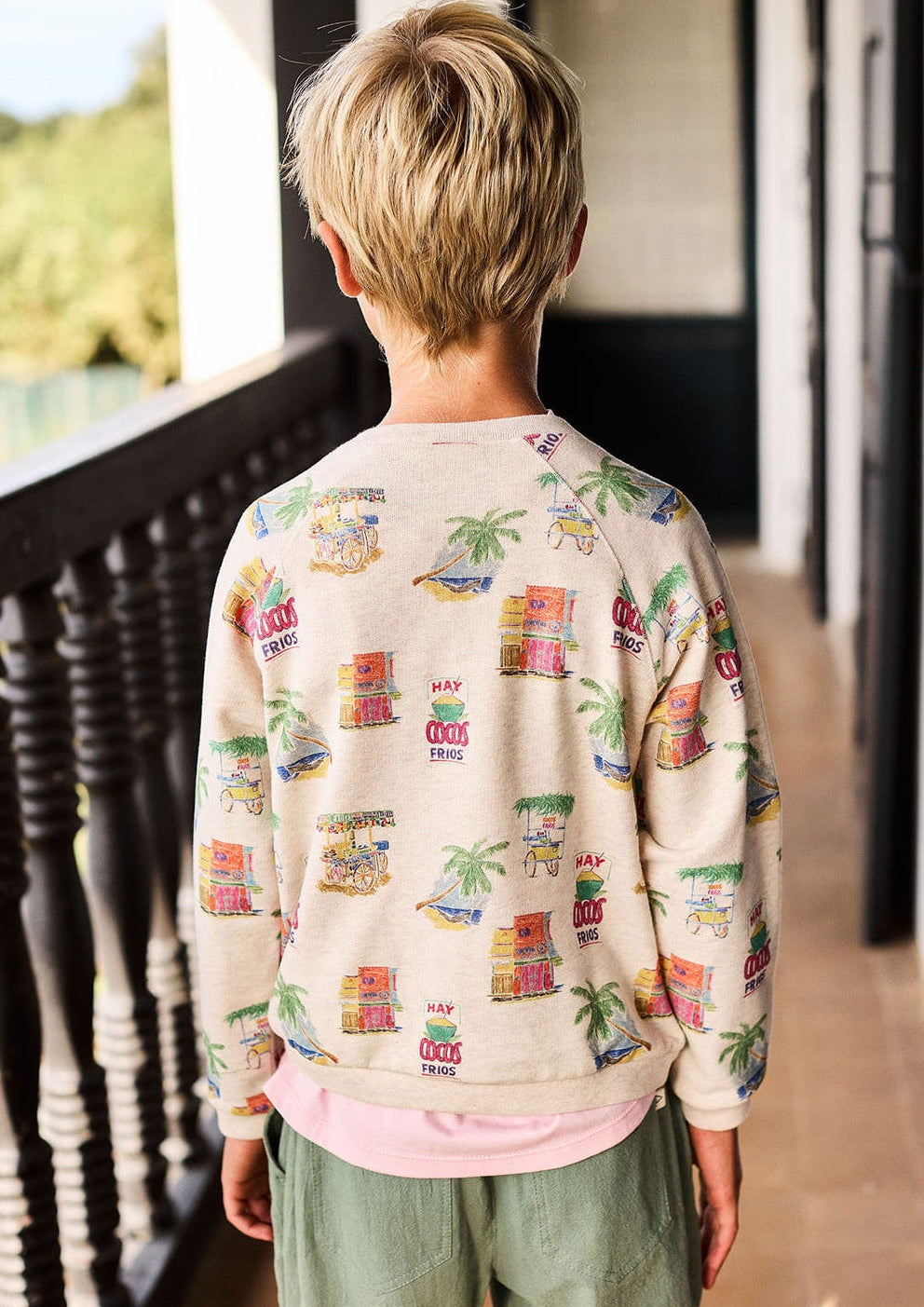 A child with short blonde hair stands on a balcony facing away, wearing the Arsene & Les Pipelettes Boys' and Girls' Mini Coco Sweatshirt with colorful prints and green pants.