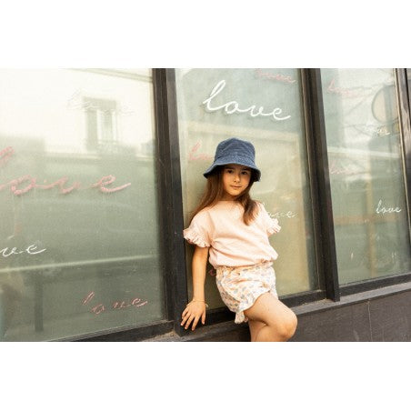 A young girl wears the Louis Louise Girls’ Naomie T-Shirt, a light-colored top with ruffled sleeves, and stands in front of a window decorated with the word “love” written multiple times.