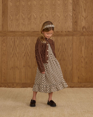 A young girl stands indoors in the Rylee & Cru Girls' Lexi Dress with ruffles and a smocked bodice, styled with a brown cardigan, loafers, and a matching headband, set against a wooden panel wall and beige carpet.