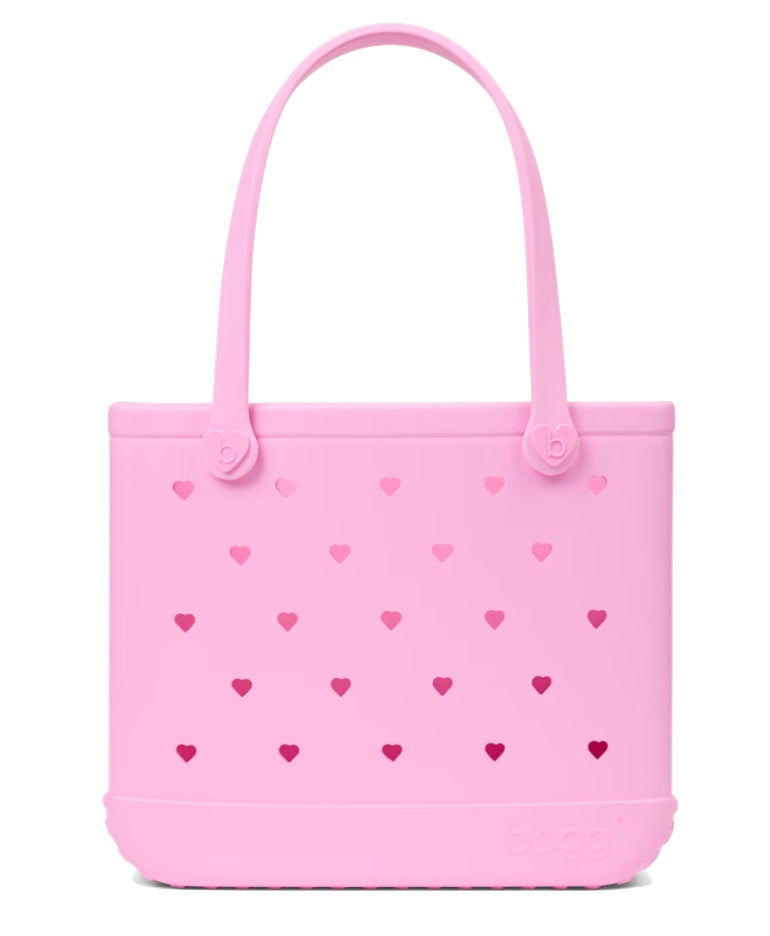 A light pink Baby Bogg® Bag by Bogg Bags, featuring small heart-shaped cutouts and two handles, crafted from a flexible, washable material and displayed against a white background.