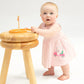 A baby girl wearing the Angel Dear Baby Girl's Bamboo Tank Dress & Bloomer in pink and white stripes stands holding a wooden stool with a small toy on top, looking slightly upward.