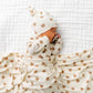 A baby lies on a white quilted blanket, dressed in the Lou Lou & Company Baby Teddy Knotted Gown with a matching hat, partially covered by the coordinating bear-print blanket—an adorable ensemble designed for easy diaper changes.