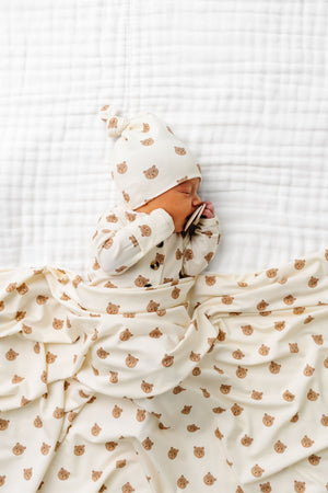 A baby lies on a white quilted blanket, dressed in the Lou Lou & Company Baby Teddy Knotted Gown with a matching hat, partially covered by the coordinating bear-print blanket—an adorable ensemble designed for easy diaper changes.