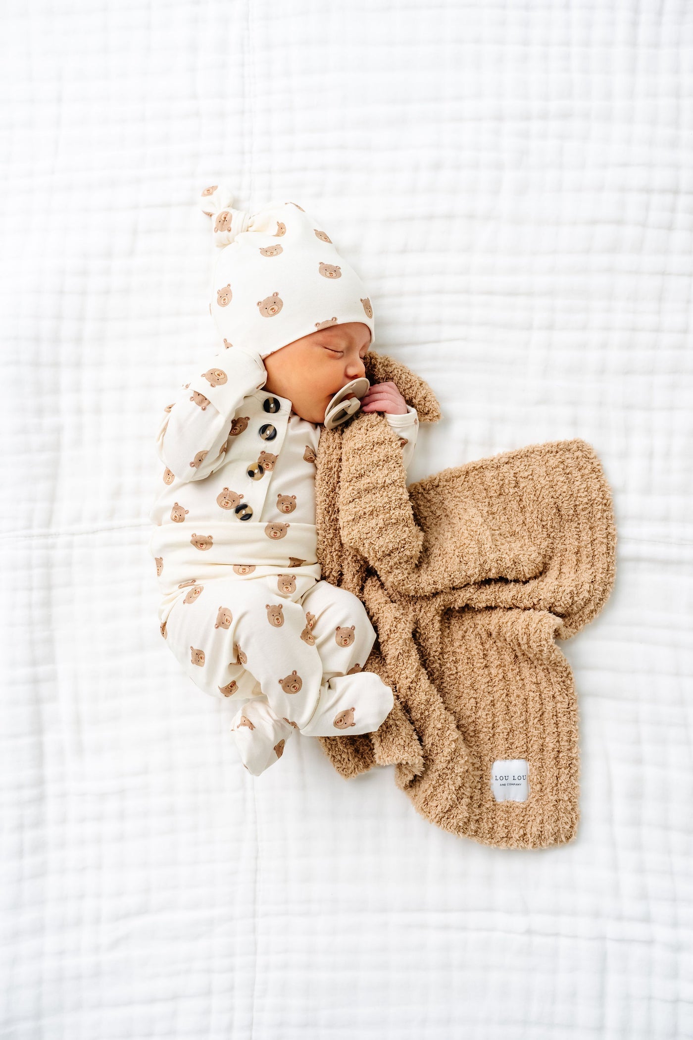 A baby dressed in Lou Lou & Company’s Baby Boys’ and Boys’ Teddy Top & Bottoms, featuring a silky-soft bear pattern with built-in footies and a matching hat, sleeps on a white blanket while holding a tan textured blanket and using a pacifier.