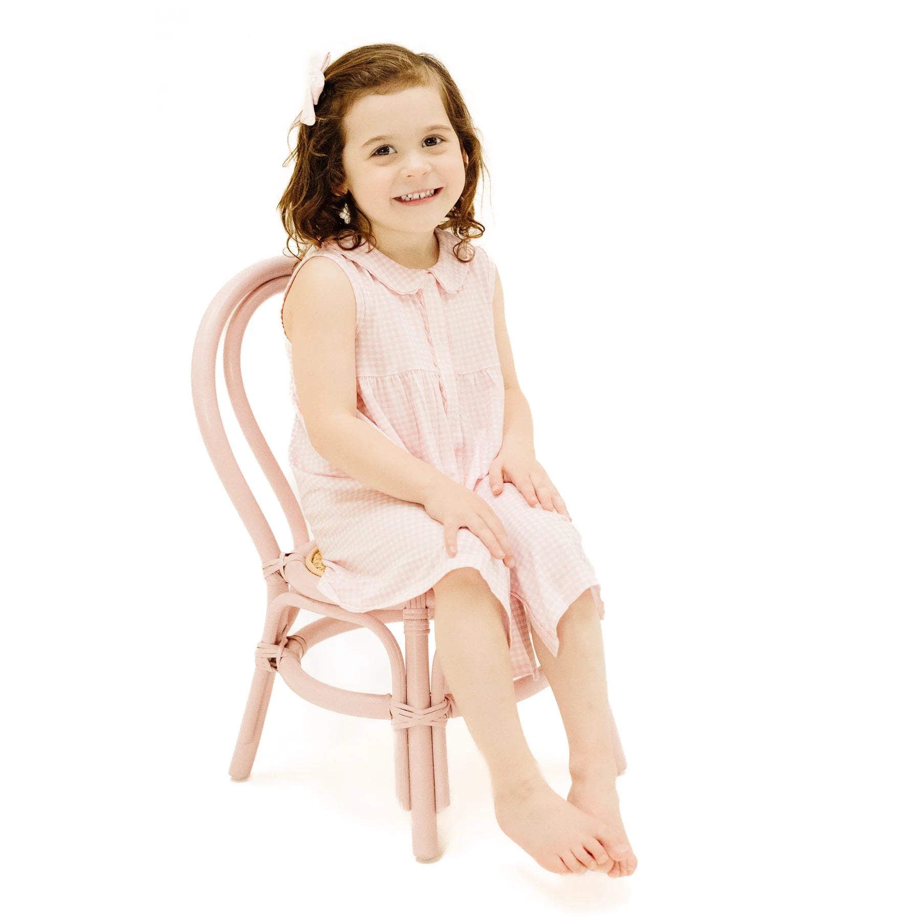 A young girl with brown hair, barefoot and wearing the Angel Dear Jill Peter Pan Button Front Dress with Ric-Rac Trim and Diaper Cover, sits on a pink chair against a white background.