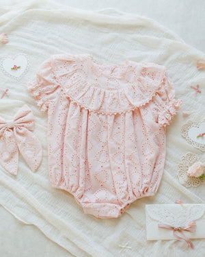 The Noralee Eva Romper by Noralee, crafted from pink heart eyelet fabric, features a frilled collar and puffed sleeves. It's laid on textured white fabric surrounded by decorative paper hearts, bows, and lace accents.