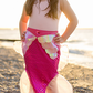 A child wearing the Great Pretenders Mermaid Glimmer Skirt, featuring holographic scales and seashell designs, paired with a tiara, smiles at the camera on a pebble beach with the ocean in the background.