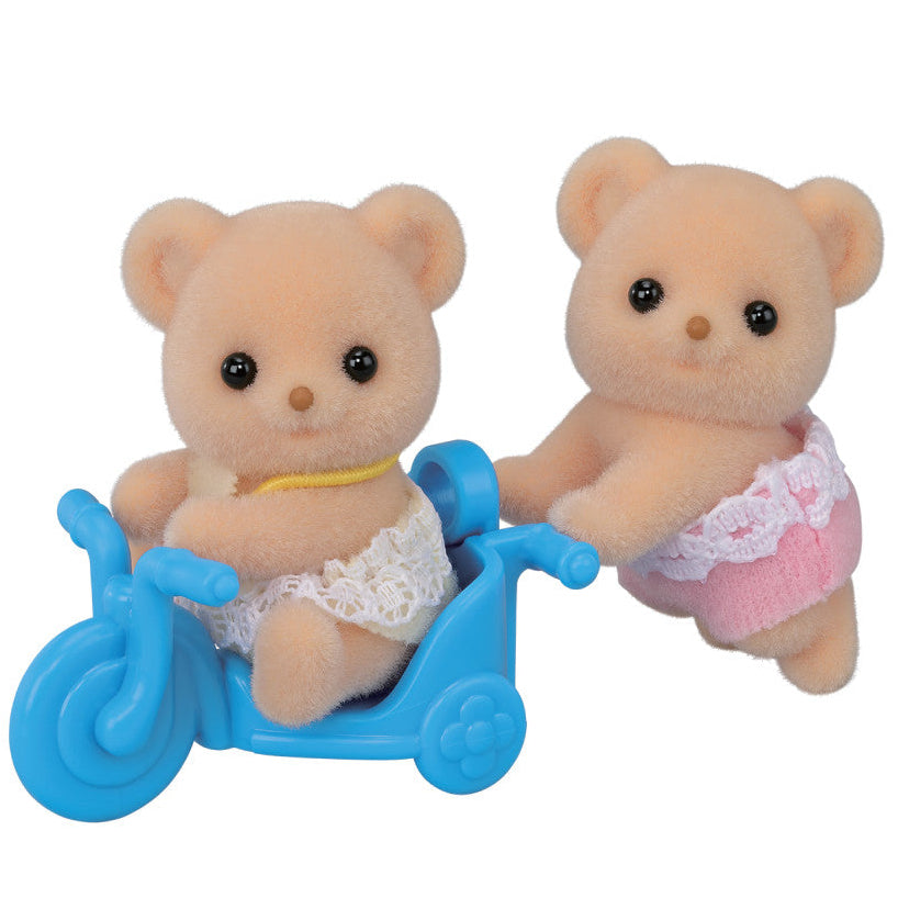 Two fuzzy Biscuit Bear Twins by Calico Critters, each wearing lace-trimmed diapers—one sitting on a blue tricycle while the other stands behind, appearing to push or guide the tricycle.