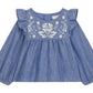 The Louise Misha Baby Girls’ Mikani Set features a blue long-sleeve cotton Oxford blouse with ruffled shoulders and white floral embroidery on the chest, shown on a white background—a charming piece from our embroidered baby outfit collection.