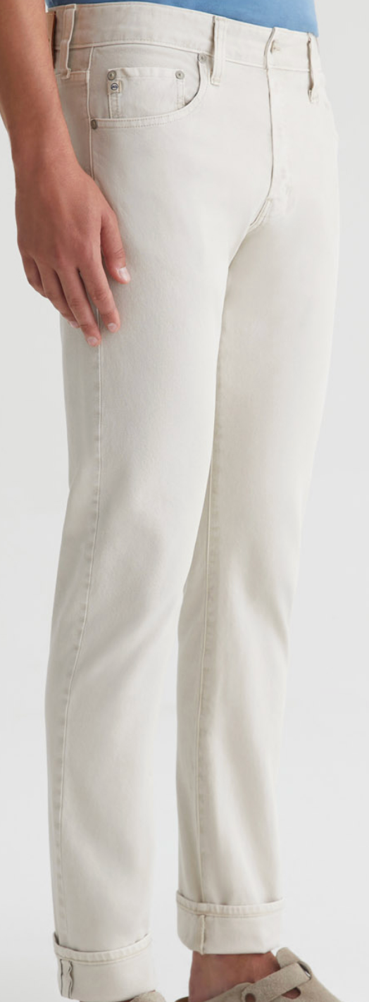 A person wears light beige Tellis Selvage jeans by AG Jeans, cuffs rolled up, shown mid-torso to feet against a plain background. Made from Japanese stretch selvage denim for both style and comfort.