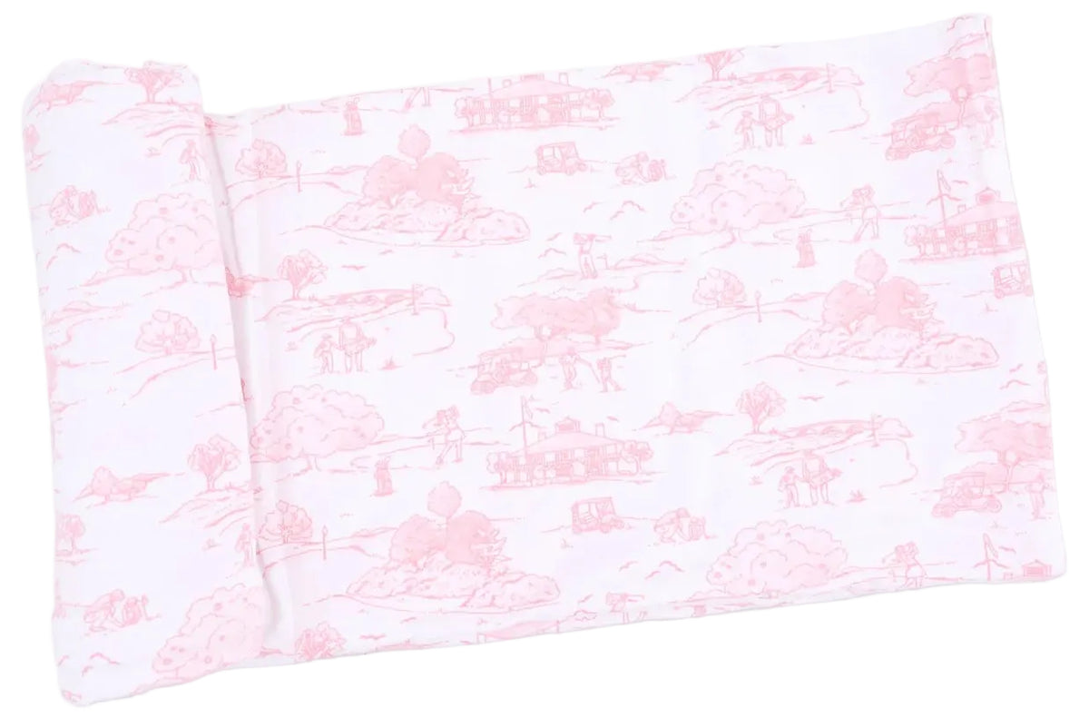 A rectangular white fabric with a light pink toile pattern depicting rural scenes—including houses, trees, and figures—is shown laid flat with one end slightly rolled. This charming piece is the Angel Dear Swaddle Blanket, perfectly soft and gentle for your baby.