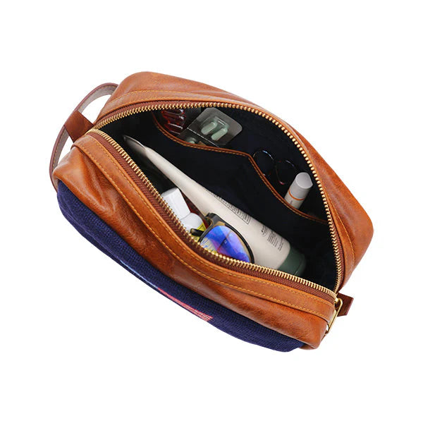 A Smathers & Branson Toiletry Bag in brown and navy Italian leather, featuring a zipper that is partially open to display contents like sunglasses, lotion, and assorted personal care items inside.