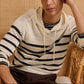 A person wearing the Onia Frank Hooded Sweater, a light-colored linen piece with black horizontal stripes, sits on a wicker chair against a woven background.