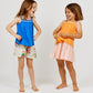 Two young girls stand barefoot, smiling and facing each other. One wears the Arsene & Les Pipelettes Girls' Solid Voile Strap Top in blue with printed shorts; the other wears an orange breathable summer top paired with a pleated pink skirt. Both wear beaded bracelets.