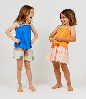 Two young girls stand barefoot, smiling and facing each other. One wears the Arsene & Les Pipelettes Girls' Solid Voile Strap Top in blue with printed shorts; the other wears an orange breathable summer top paired with a pleated pink skirt. Both wear beaded bracelets.